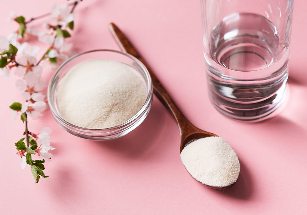 A glass bowl and wooden spoon hold white powder beside a clear glass of water on a pink surface, accompanied by small white cherry blossoms.